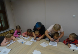 Sześcioro dzieci siedzi przy stole. pani Agnieszka pochylona jest nad dzieckiem, któremu pomaga podczas malowania.